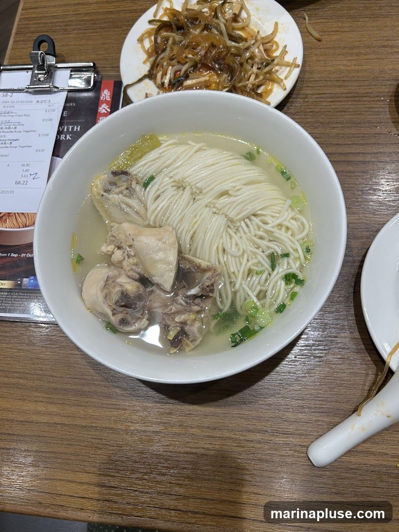 A bowl of chicken noodle soup with noodles, chicken pieces, and green onions, served on a wooden table. A small plate of pickled vegetables is in the background. A receipt is clipped to a stand next to the bowl.