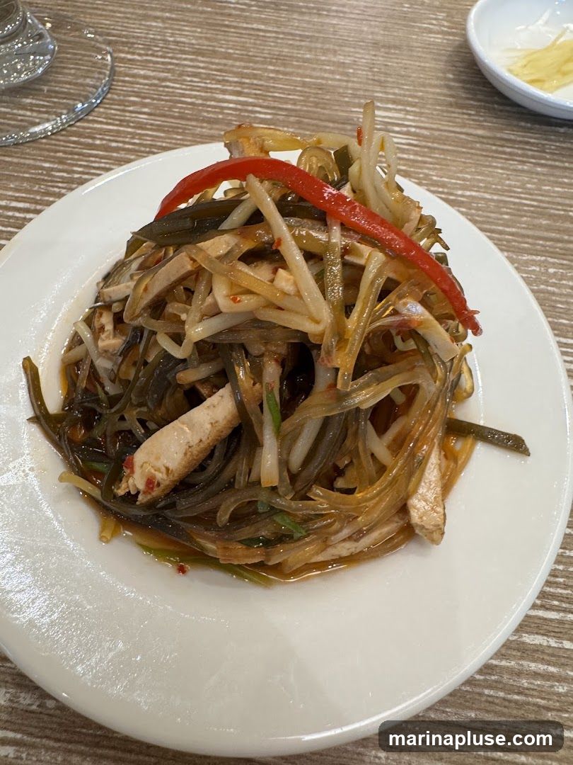 A white plate holds a salad composed of thin noodles, strips of tofu, and dark green seaweed, all tossed in a light-colored sauce. A slice of red bell pepper is visible on top. The salad is served on a wooden table, with a glass and a small white dish in the background.