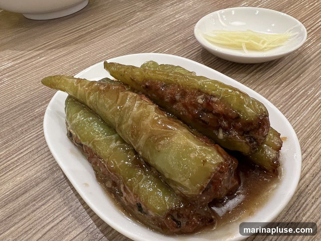 A plate of stuffed green peppers in a savory sauce, served with a side of thinly sliced ginger. The peppers appear to be filled with a meat mixture and are glistening in the sauce. The dish is presented on a wooden table.