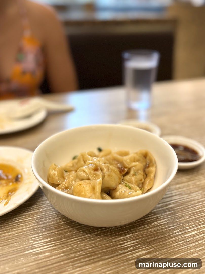 A close-up shot of a bowl of dumplings in a light brown sauce, garnished with green onions. A small dish of dark dipping sauce is visible to the right. In the background, a person wearing an orange and pink floral top is seated at a table with a glass of water. The table has a wood-grain texture.