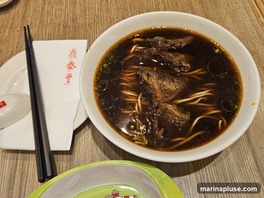 A bowl of beef noodle soup with large pieces of braised beef, noodles, and green onions in a dark broth. A napkin with red Chinese characters and a pair of chopsticks are on the side.