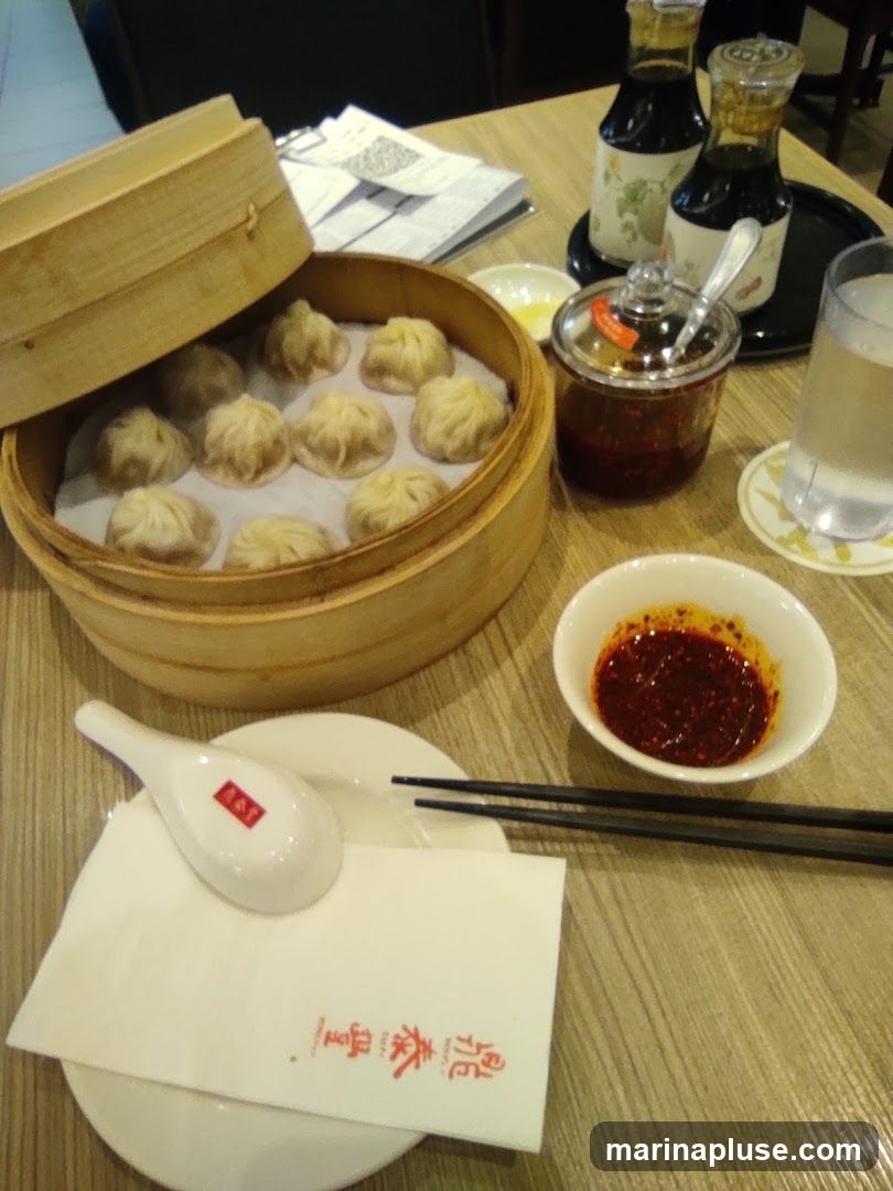 A bamboo steamer filled with dim sum is placed on a wooden table. Next to it are small bowls of chili oil and pickled ginger, two bottles of soy sauce, a glass of water, chopsticks, and a spoon with a napkin. The napkin has red Chinese characters printed on it.