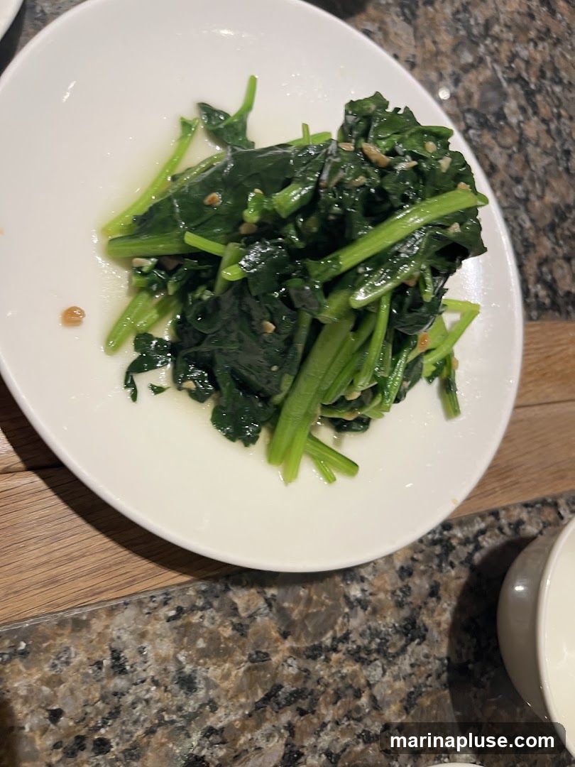 A white plate filled with sautéed spinach, garnished with minced garlic. The plate is on a granite countertop next to a wooden surface.