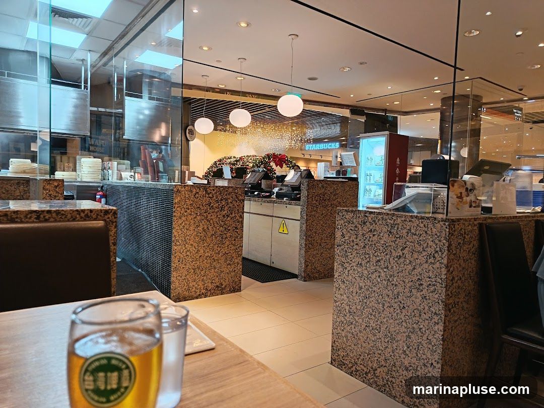 The image shows the interior of a restaurant with a focus on the counter area and seating. In the foreground, a table with a glass of beer and a glass of water is visible. Behind the table, a counter with a speckled granite finish is prominent. Further back, a service counter with a cash register and a refrigerator is seen. The kitchen area is visible through glass partitions, with stacks of steamers and stainless steel equipment. A sign for "STARBUCKS" is visible in the background, suggesting the restaurant is located within a mall or shopping center. Festive decorations, possibly for Christmas, are also present.