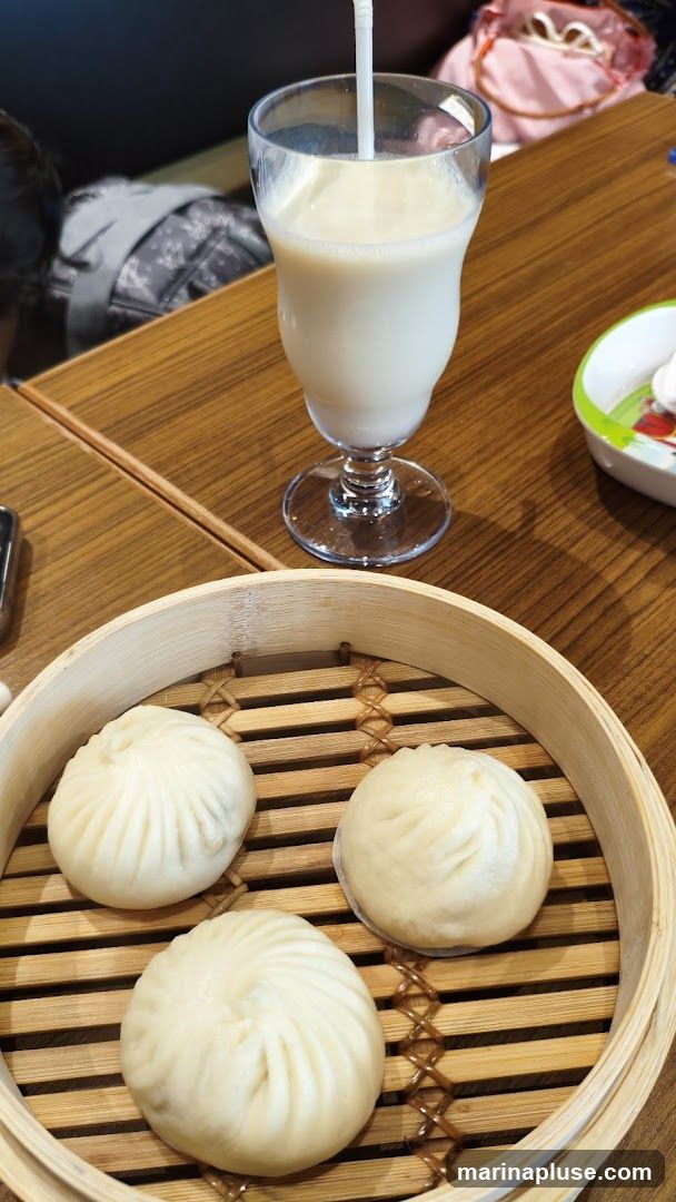 A bamboo steamer basket holds three steamed buns. A glass of a light-colored beverage with a straw sits on a wooden table. In the background, a person is seated, and a pink bag is visible.