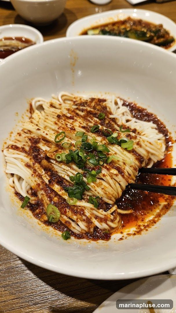 A close-up shot of a bowl of noodles with a spicy sauce and chopped green onions. Chopsticks are visible, holding a portion of the noodles. In the background, other dishes are partially visible.