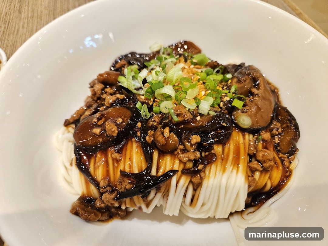 A close-up shot of a bowl of noodles topped with a dark sauce, minced meat, mushrooms, and chopped green onions.