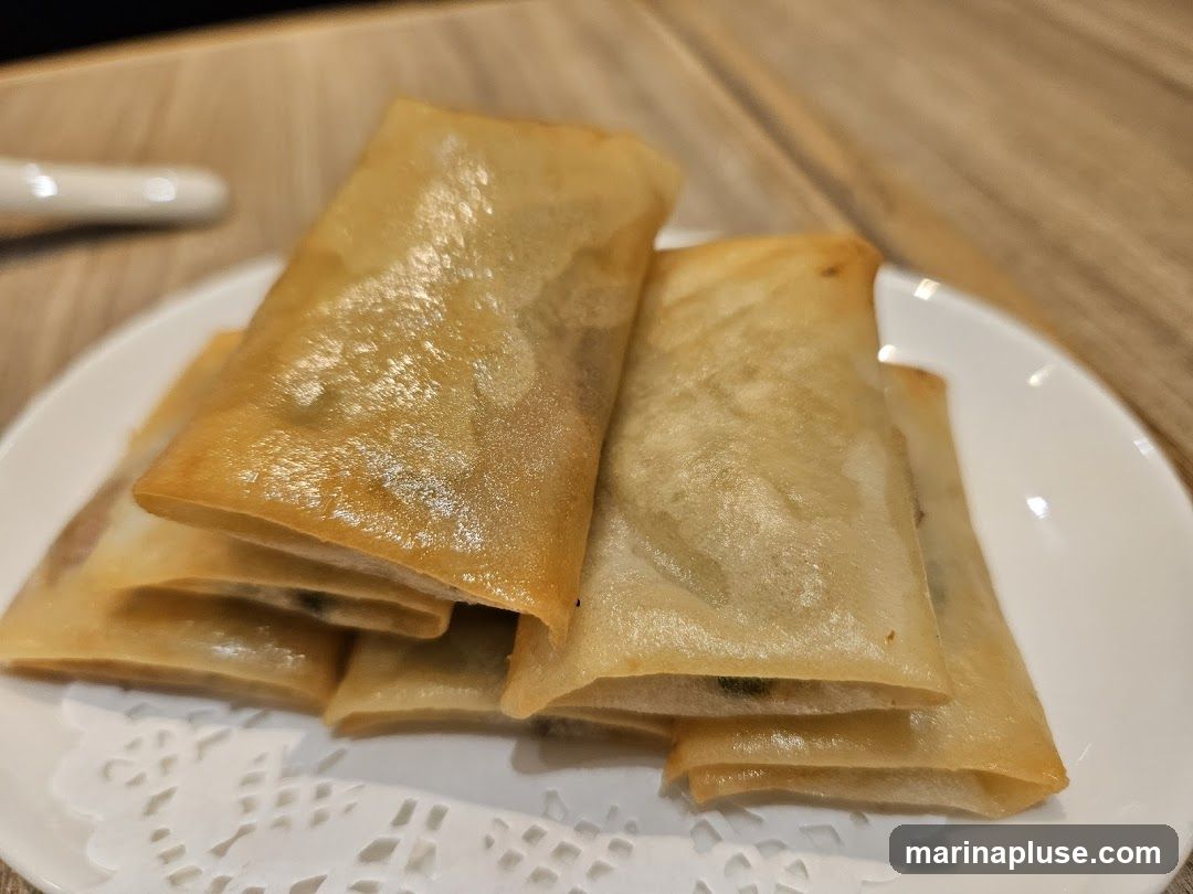 A close-up shot of several golden-brown spring rolls stacked on a white plate with a decorative paper liner. The spring rolls appear to be fried and have a crispy, translucent wrapper.