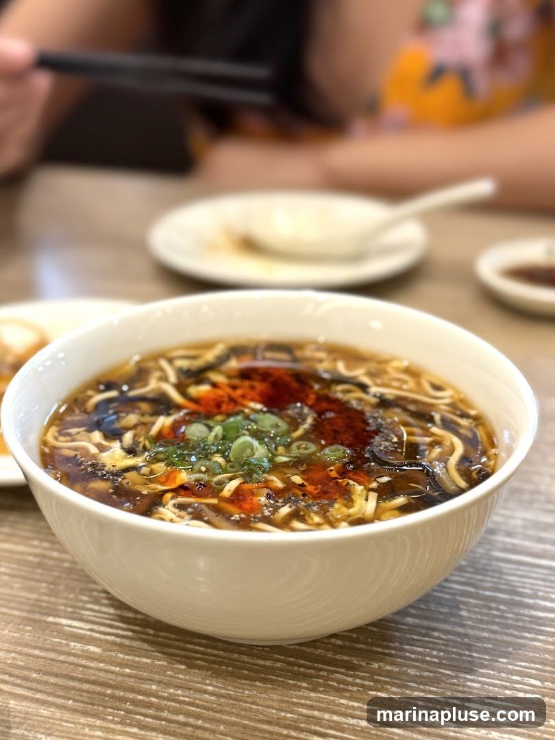 A close-up shot of a bowl of hot and sour soup, garnished with chopped green onions and chili oil. In the background, two people are seated at a table, with one person holding chopsticks.