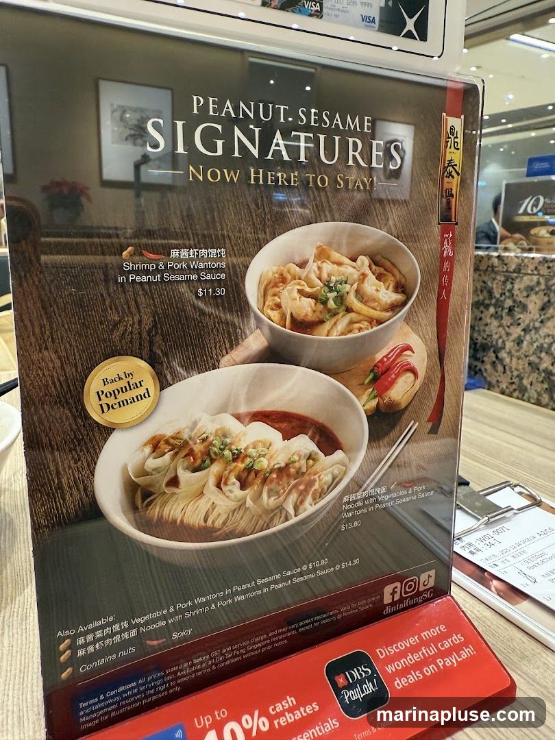 A menu board showcasing "Peanut Sesame Signatures" with images of wontons. It highlights "Shrimp & Pork Wantons in Peanut Sesame Sauce" for $11.30 and "Vegetable & Pork Wantons in Peanut Sesame Sauce" for $10.80. Other options include "Noodle with Vegetables & Pork Wantons in Peanut Sesame Sauce" for $13.80 and "Noodle with Shrimp & Pork Wantons in Peanut Sesame Sauce" for $14.30. It also notes that the dishes contain nuts and are spicy. A promotional section for DBS PayLah! is visible at the bottom.