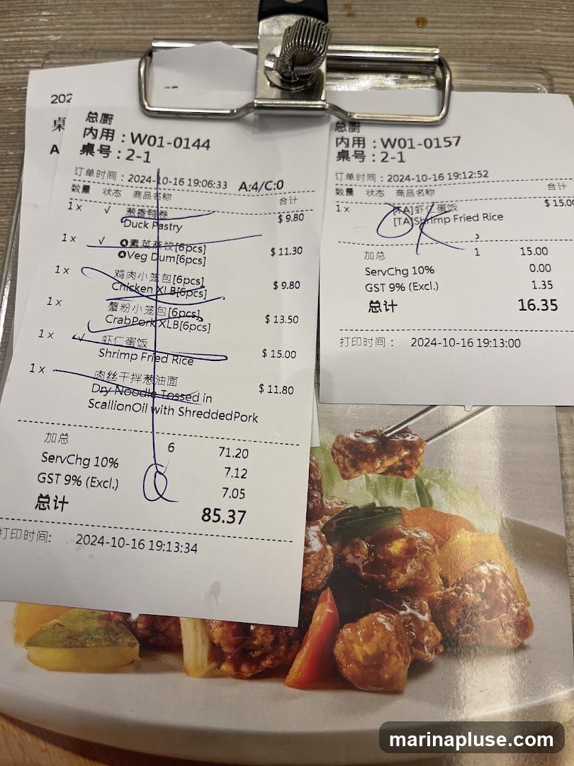 Two receipts are clipped together, with a plate of food visible below. The receipts detail various food orders and their prices, including items like 'Duck Pastry', 'Veg Dum', 'Chicken XLB', 'Crab Pork XLB', 'Shrimp Fried Rice', and 'Dry Noodle Tossed in Scallion Oil with Shredded Pork'. Service charges and GST are also listed. The food appears to be a Chinese dish with meat and vegetables.