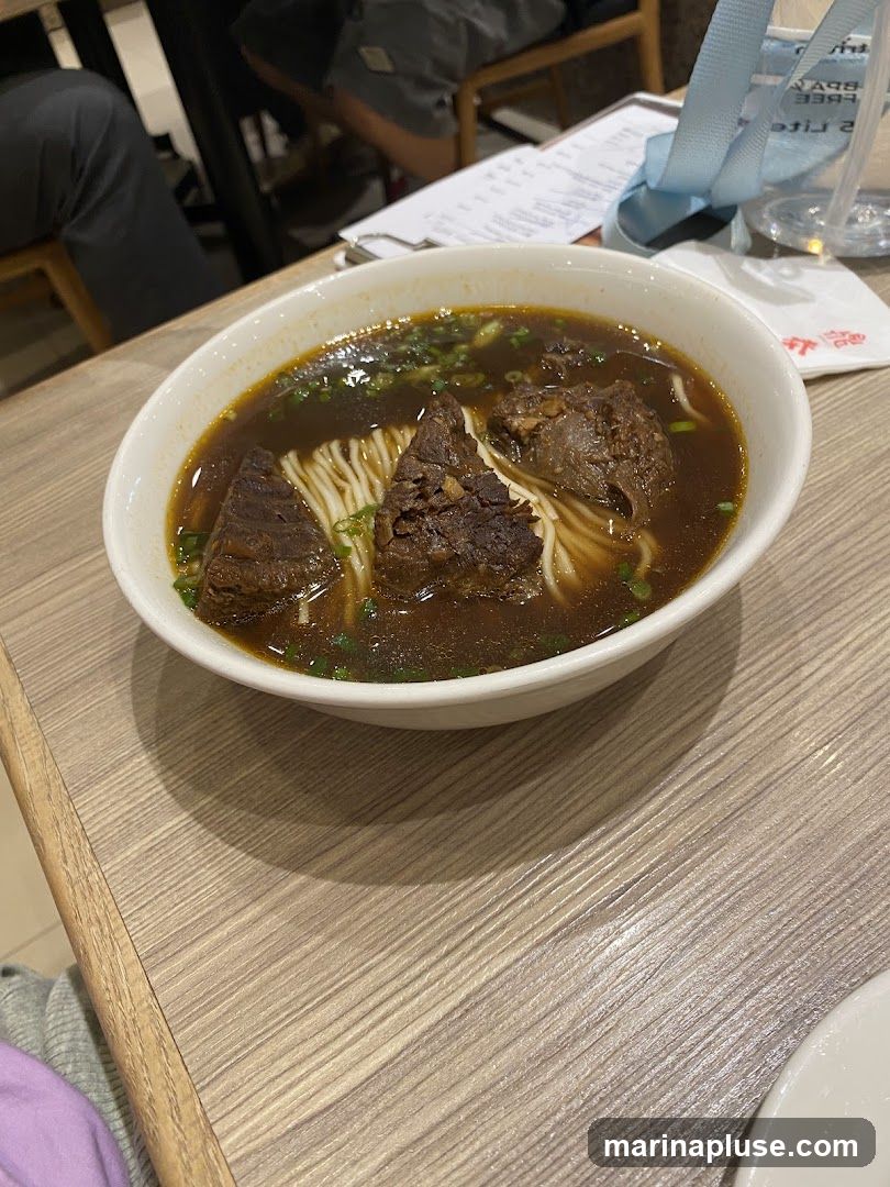 A close-up shot of a bowl of beef noodle soup. The soup is a dark broth with thick noodles and large chunks of braised beef. Chopped green onions are sprinkled on top. The bowl is placed on a light-colored wooden table. In the background, a menu and a clear plastic cup with a blue strap are visible.