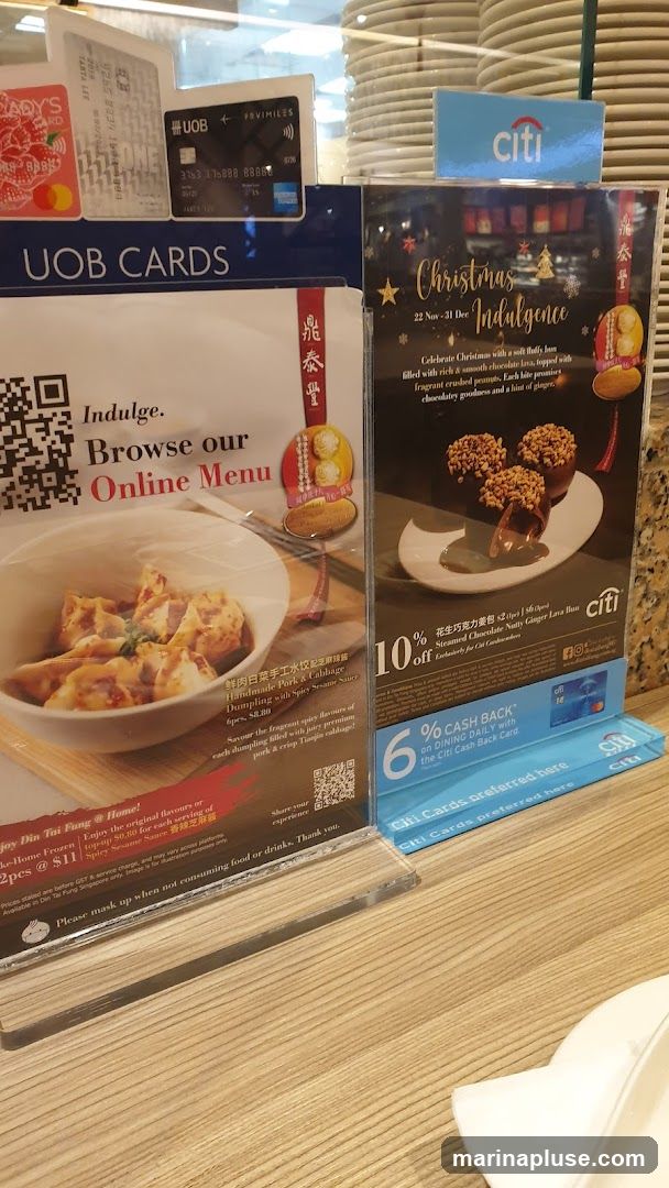 Two promotional displays are shown on a wooden counter. The left display, titled "UOB CARDS Indulge. Browse our Online Menu," features an image of dumplings and a QR code. It also mentions "Din Tai Fung @ Home!" and offers "Handmade Pork & Cabbage Dumpling with Spicy Sesame Sauce, 6pcs @ $11." The right display advertises "Christmas Indulgence" with a chocolate dessert and offers "10% off Steamed Chocolate Nutty Ginger Lava Bun" exclusively for Citi Cardholders. It also highlights "6% CASH BACK on DINING DAILY with the Citi Cash Back Card." Several credit cards, including UOB and Citi, are visible.