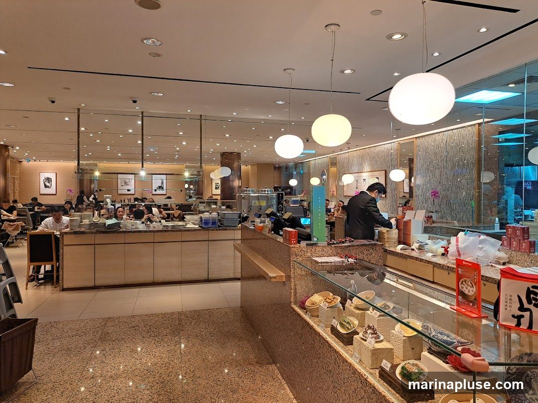 An interior view of a busy restaurant with diners seated at tables and a food display counter in the foreground. A staff member in a suit and mask is visible behind the counter, preparing food. The display case showcases various small dishes and food items. The restaurant has modern lighting and decor.
