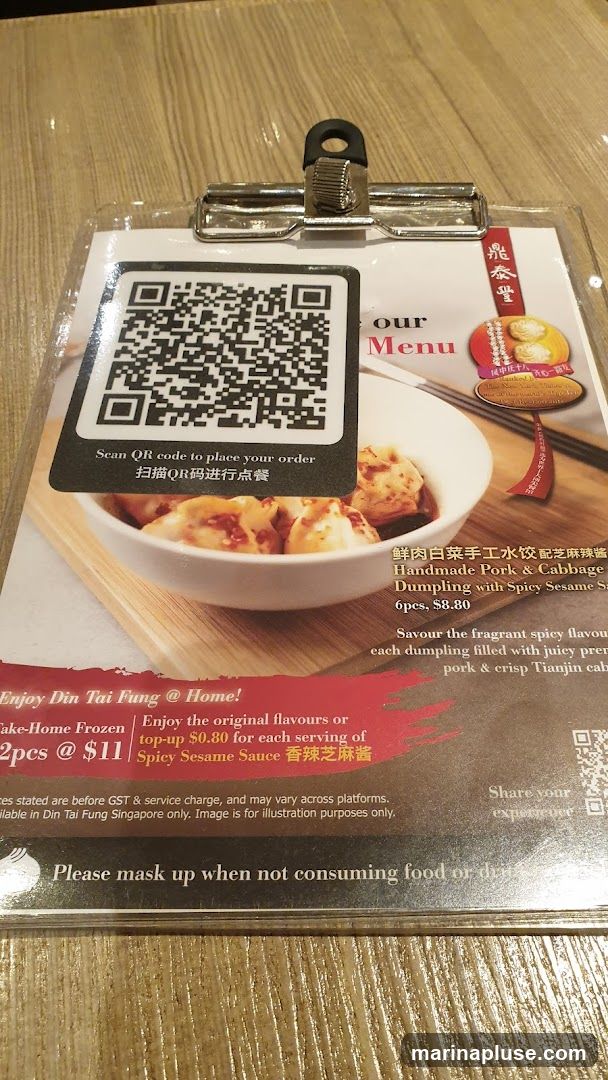 A menu featuring a QR code for ordering, with an image of dumplings. It highlights "Handmade Pork & Cabbage Dumpling with Spicy Sesame Sauce" for 6pcs at $8.80. Also offers "Take-Home Frozen" dumplings for 2pcs at $11 and a top-up for Spicy Sesame Sauce. Includes a note about masking up when not consuming food or drink.