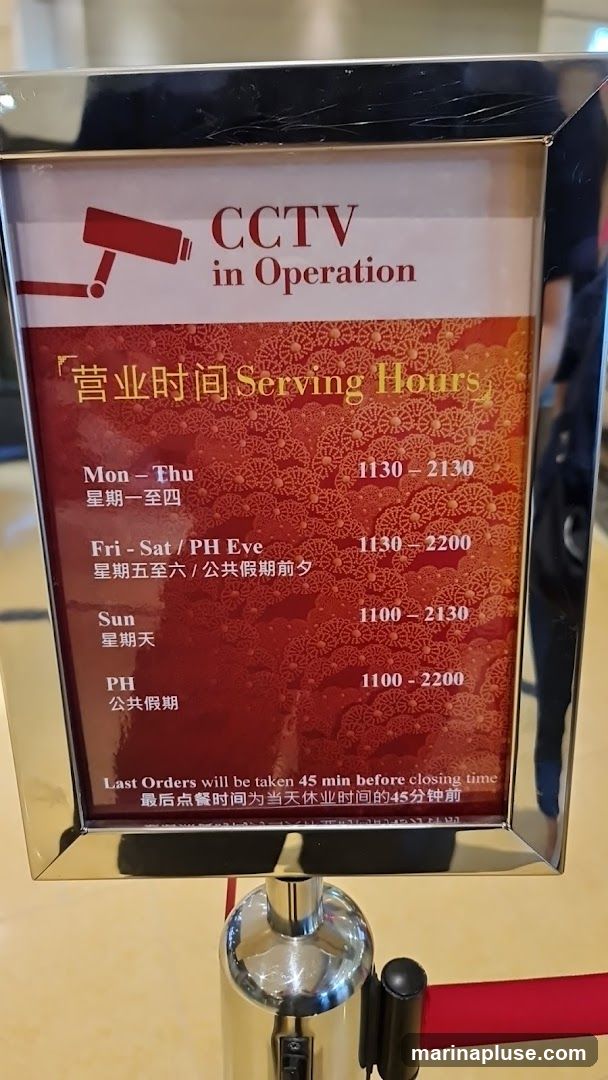 A sign displays the serving hours for a business, indicating that CCTV is in operation. The hours are listed for Monday-Thursday (11:30-21:30), Friday-Saturday/PH Eve (11:30-22:00), Sunday (11:00-21:30), and PH (11:00-22:00). It also states that last orders are taken 45 minutes before closing time.