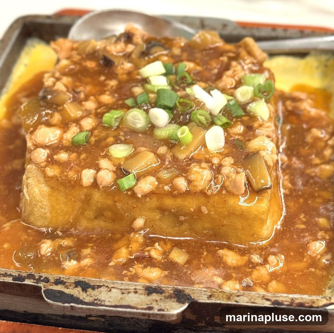 A close-up shot of a square piece of fried tofu served on a sizzling platter. The tofu is covered in a savory brown sauce with minced meat, mushrooms, and chopped green onions. The dish appears to be a hot and hearty meal.