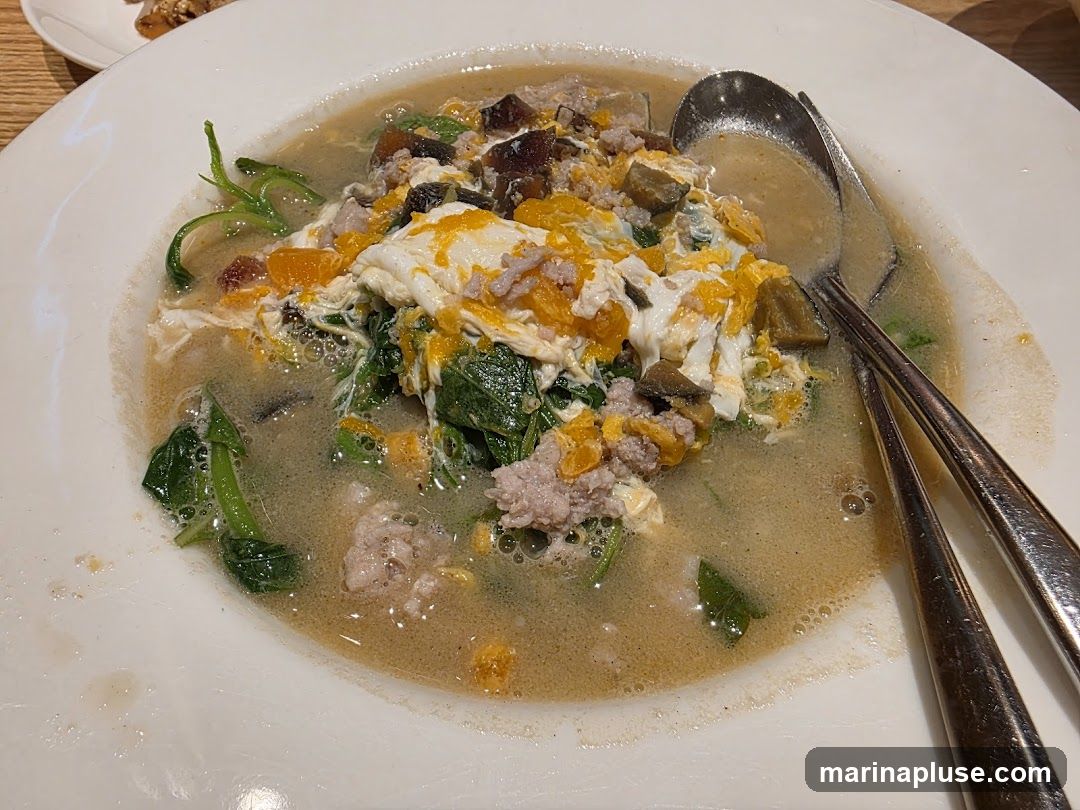 A close-up shot of a bowl of soup with minced pork, scrambled eggs, and leafy green vegetables. Two spoons are resting in the bowl.