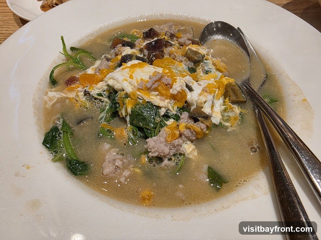A close-up shot of a bowl of soup with minced pork, scrambled eggs, and leafy green vegetables. Two spoons are resting in the bowl.