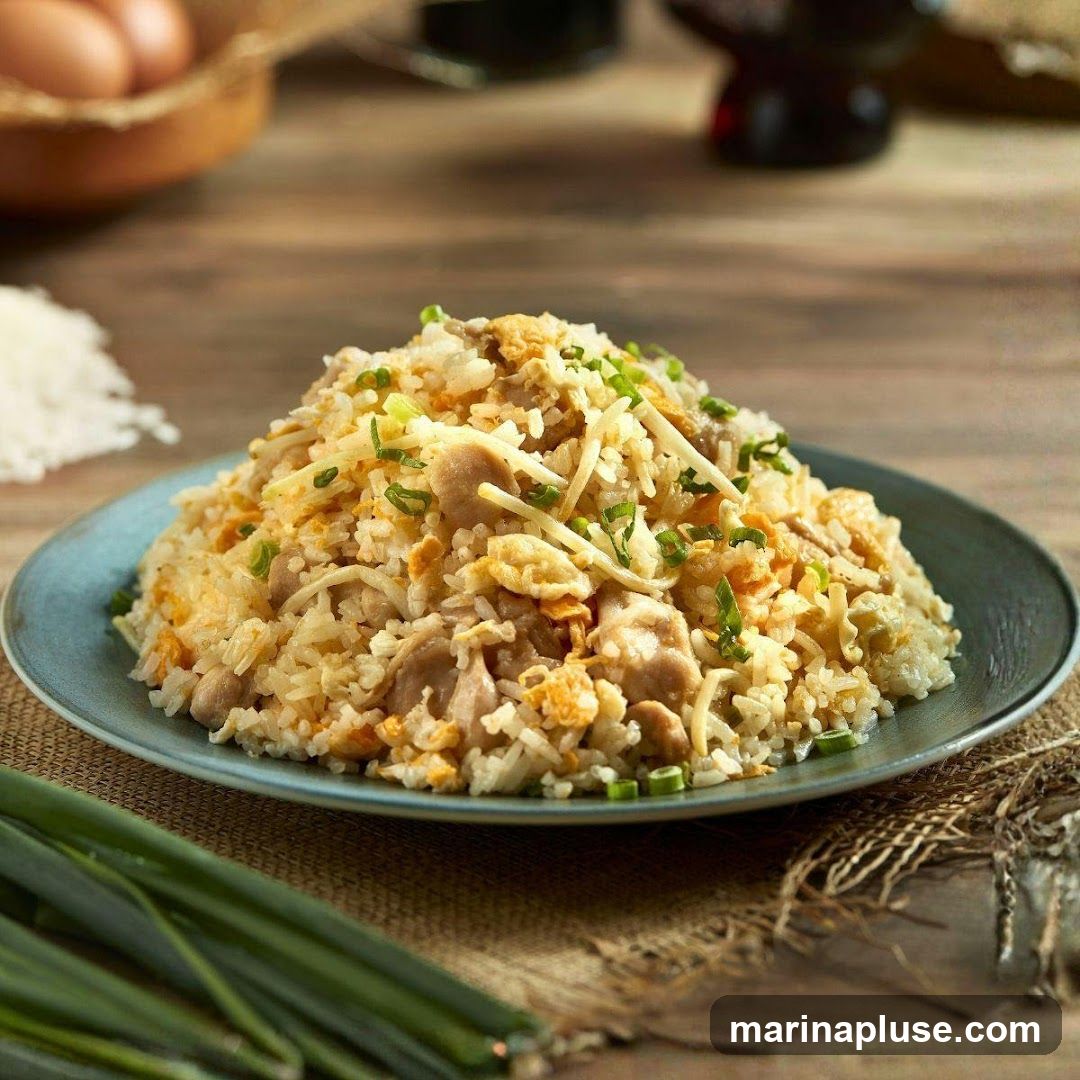 A plate of chicken fried rice garnished with chopped green onions. In the background, there are eggs in a basket, a pile of uncooked rice, and two bottles of dark liquid. The food is presented on a wooden table with a burlap placemat.