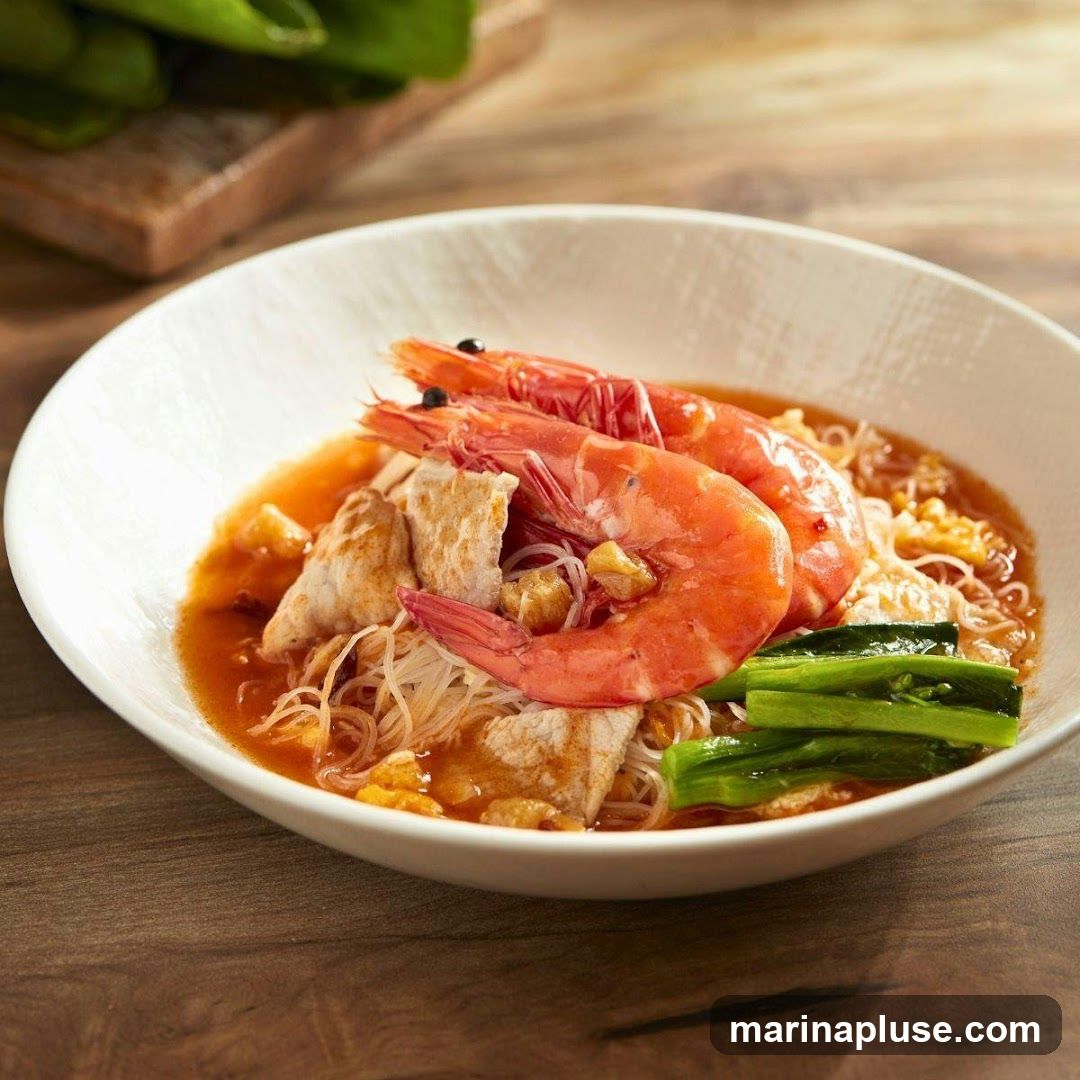 A close-up shot of a bowl of noodle soup with two large shrimp, pieces of fish, and green vegetables. The soup has a reddish-orange broth and is served in a white bowl on a wooden surface.