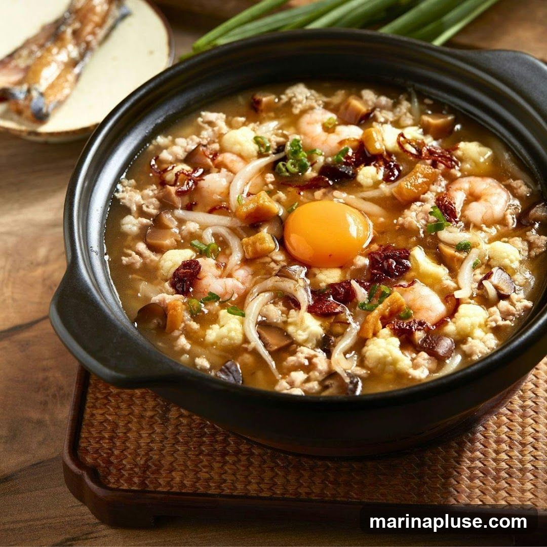 A close-up shot of a steaming hot stew in a dark ceramic pot, placed on a woven mat. The stew is rich with ingredients including minced meat, plump shrimp, cauliflower florets, mushrooms, and noodles. A bright, glistening egg yolk sits prominently in the center, surrounded by chopped scallions and other garnishes. In the blurred background, a plate holds what appears to be grilled fish.