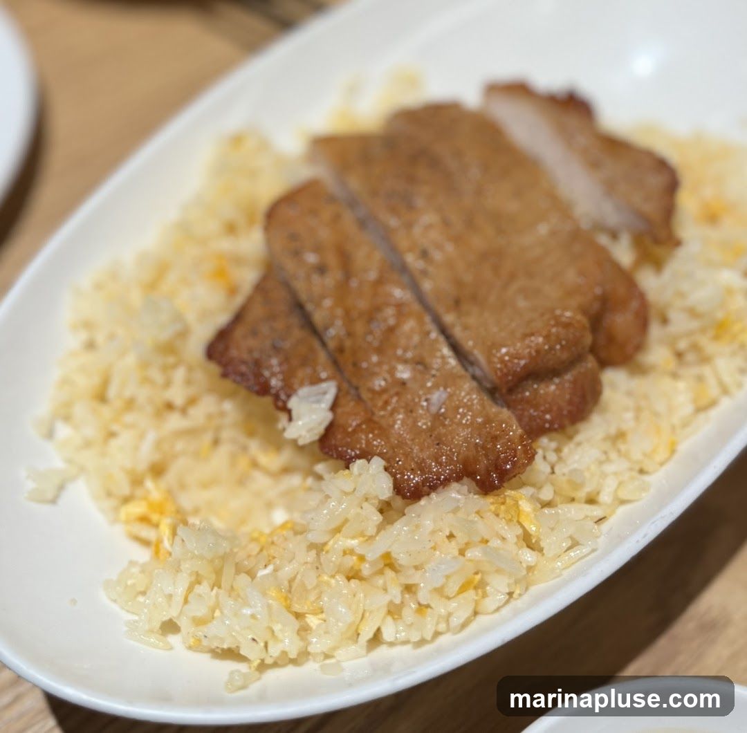 A close-up shot of a white oval plate filled with fried rice and topped with sliced pork chops. The fried rice appears to have scrambled egg mixed in. The pork chops are golden brown and appear to be seasoned.
