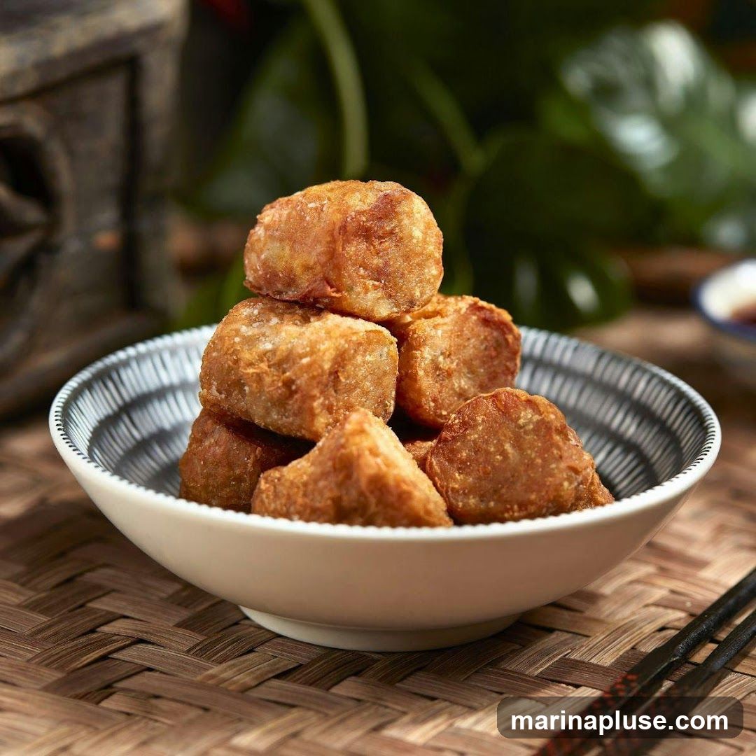 A close-up shot of a bowl filled with golden-brown fried rolls, stacked on top of each other. The bowl has a decorative rim with black and white patterns. The rolls appear crispy and are arranged in a pyramid shape. In the background, a blurred green plant and a dark, ornate object are visible. Chopsticks rest on a woven placemat in the foreground.