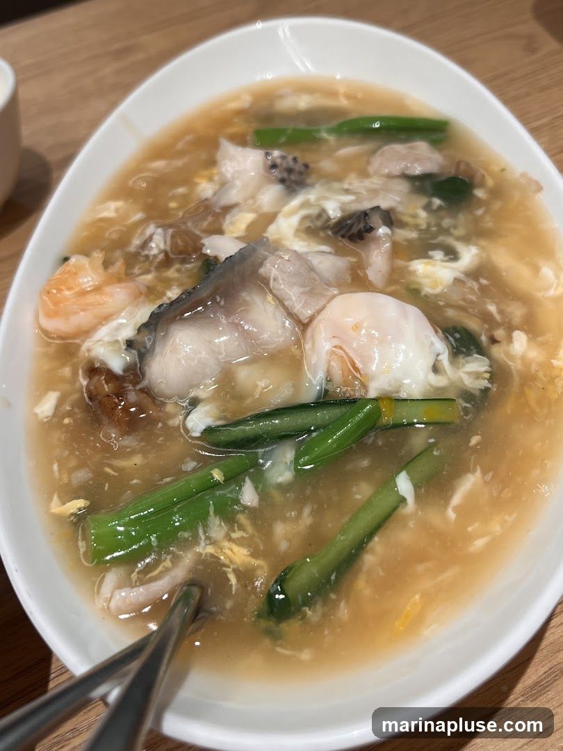 A close-up shot of a bowl of seafood noodles. The dish features a thick, savory broth with pieces of fish, shrimp, and green vegetables. A poached egg is also visible in the bowl. Chopsticks are resting in the dish.