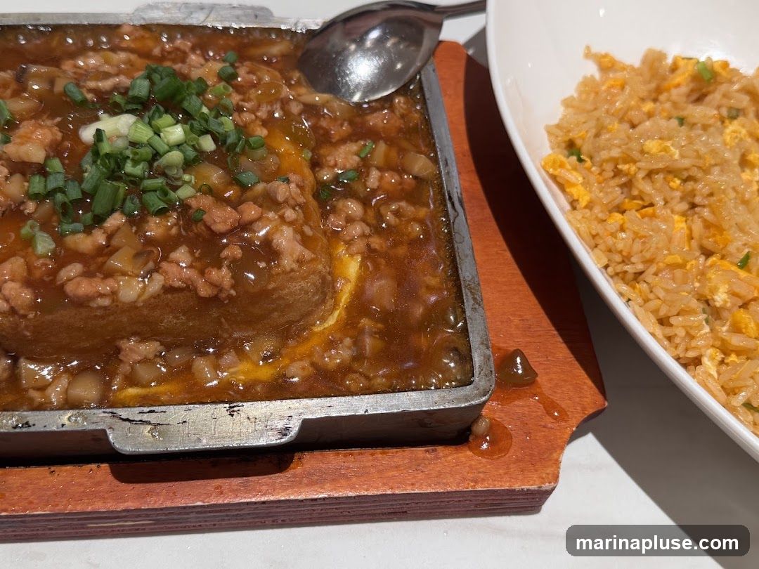 A close-up shot of a sizzling dish of tofu topped with a savory sauce and minced pork, garnished with chopped green onions. The dish is served in a metal tray on a wooden base. To the right, a bowl of fluffy fried rice with visible egg pieces is presented.