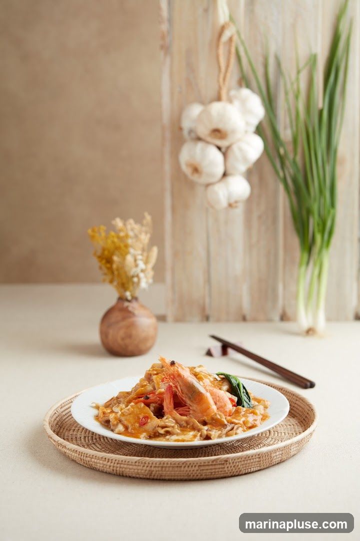 A plate of seafood noodles with large shrimp, served on a woven placemat. In the background, a bunch of garlic and green onions hang on a wooden wall, with a small vase of dried flowers to the left.