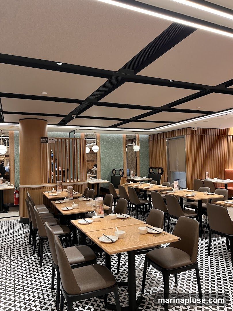 A well-lit restaurant interior with a modern design. The space features numerous tables set for dining, each with placemats, bowls, and chopsticks. The flooring has a distinctive black and white geometric pattern. The ceiling is a grid of white panels with black accents and integrated lighting. Wooden elements are used for partitions and wall paneling, creating a warm ambiance. Several chairs are visible around the tables.