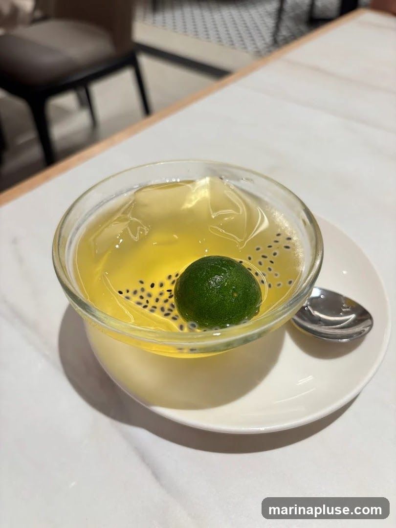 A close-up shot of a dessert in a clear glass bowl, served on a white saucer with a spoon. The dessert appears to be a yellow jelly or pudding topped with ice cubes, black seeds, and a small green lime. The background shows a table and chairs, suggesting an indoor dining setting.