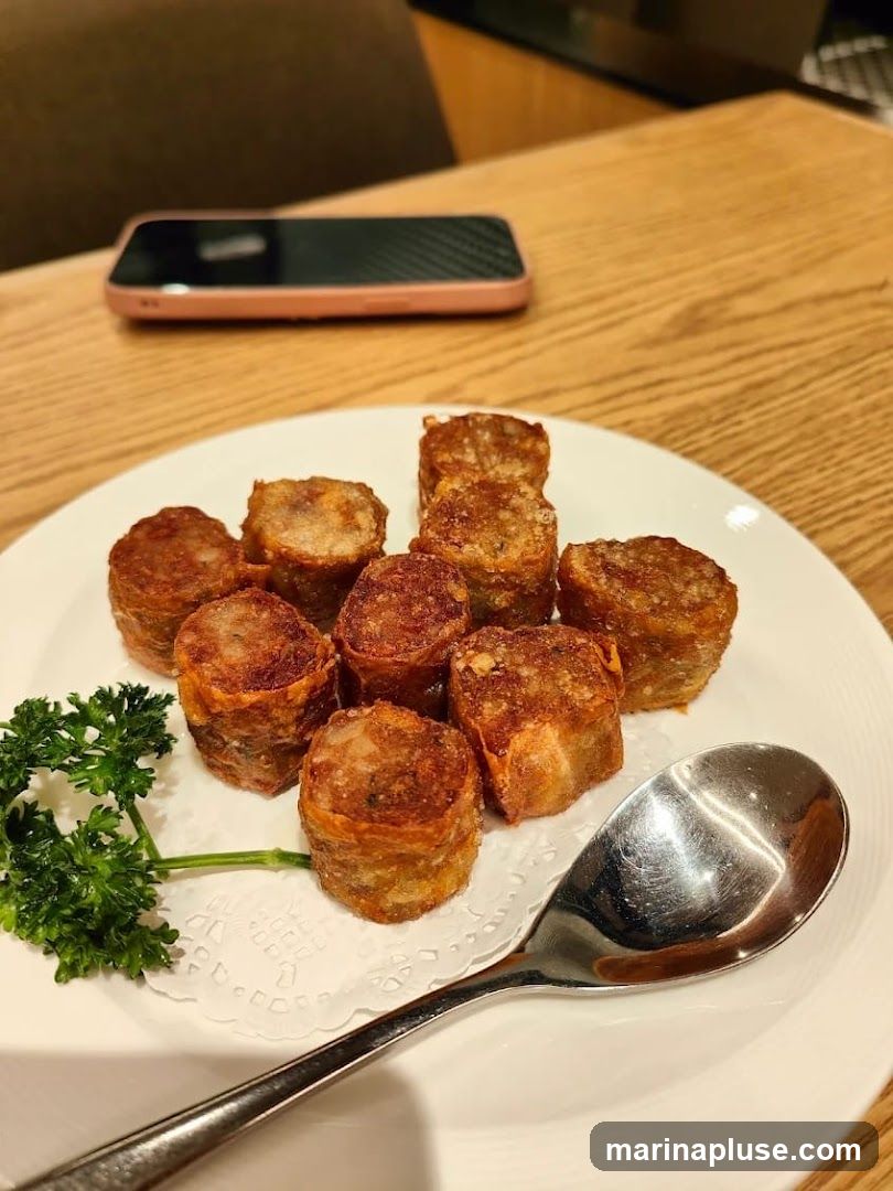A plate of fried, cylindrical food items, possibly spring rolls or dim sum, is presented on a white plate with a doily and a sprig of parsley. A spoon rests on the right side of the plate. In the background, a smartphone with a carbon fiber case is placed on a wooden table.