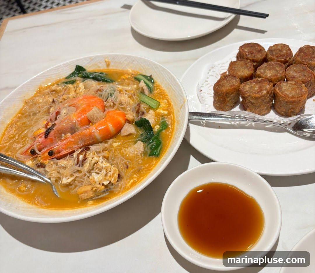 A close-up shot of a meal featuring a bowl of noodles with shrimp and vegetables, a plate of fried rolls, and a small dish of dipping sauce. The noodles appear to be thin vermicelli in a savory broth, topped with two large prawns, scrambled egg, and leafy greens. The fried rolls are cylindrical and golden brown.