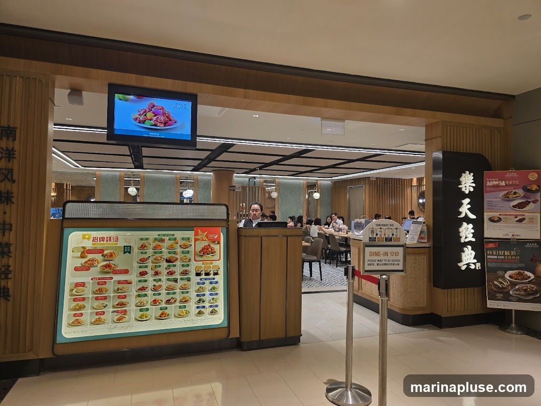 A view of a restaurant entrance with a large illuminated menu board displaying various dishes. A screen above shows a close-up of a food item. In the background, diners are seated at tables. A vertical sign with Chinese characters is visible on the right side, along with promotional posters.