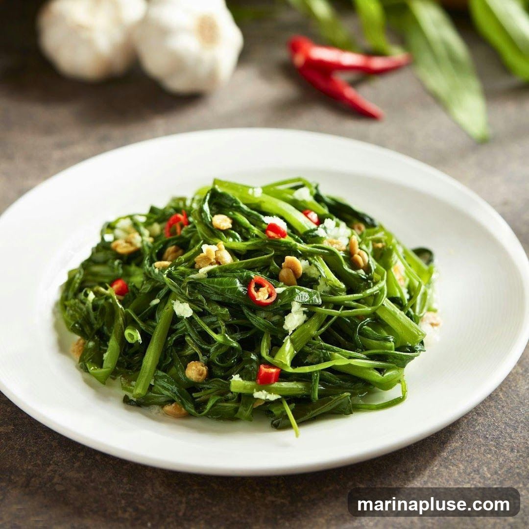 A close-up shot of a plate of stir-fried water spinach, also known as kangkong. The dish is garnished with chopped garlic, chili slices, and peanuts. In the background, out of focus, are garlic bulbs and red chilies.