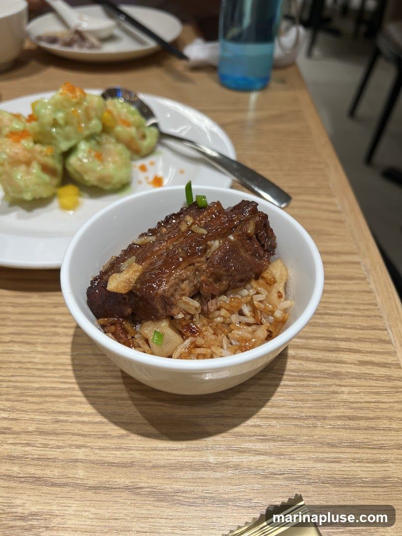 A close-up shot of a bowl of braised pork belly served over rice. The pork belly is dark brown and glistening, with visible layers of fat and meat. It is garnished with chopped green onions. The rice underneath appears to be seasoned and mixed with small pieces of something, possibly tofu or vegetables. In the background, a plate of green dumplings topped with orange roe is visible, along with cutlery and a blue water bottle. The setting appears to be a restaurant table with a light wood grain surface.