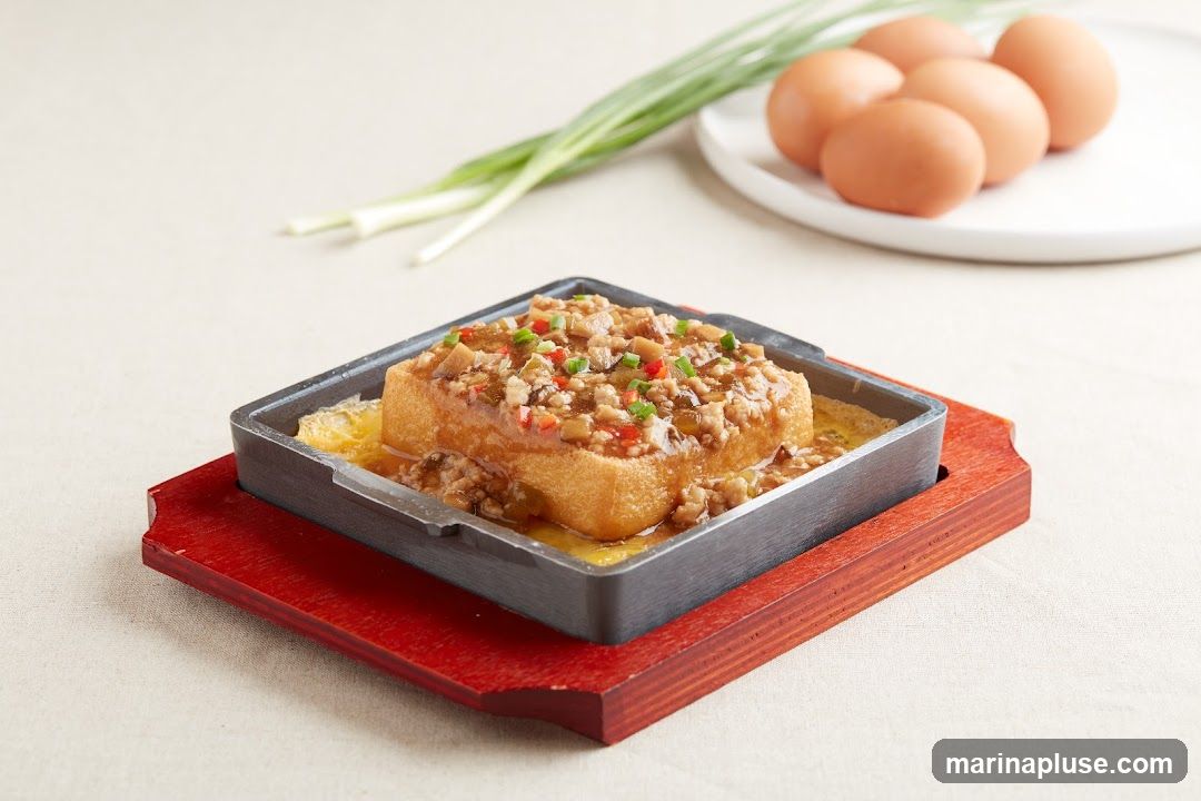 A close-up shot of a square piece of fried tofu, topped with a savory minced meat and vegetable sauce, served in a sizzling metal dish on a red wooden base. In the background, a plate of brown eggs and a bunch of green onions are visible. The dish is presented on a light-colored tablecloth.
