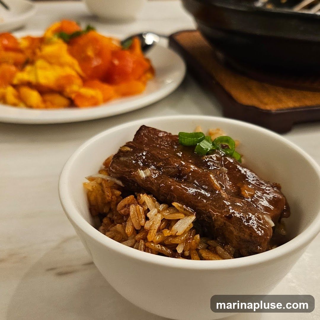 A close-up shot of a bowl of rice topped with braised beef and garnished with chopped green onions. In the background, a plate of orange and yellow food, possibly scrambled eggs with tomatoes, is visible.