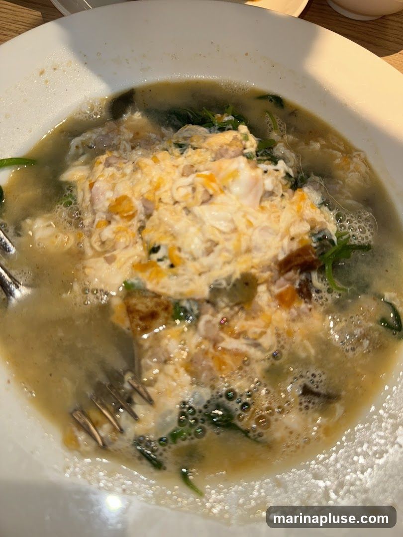A close-up shot of a bowl of soup with a topping of scrambled egg and green vegetables. A fork is visible in the lower left corner of the bowl. The soup appears to be light in color with some darker pieces and green leafy vegetables.