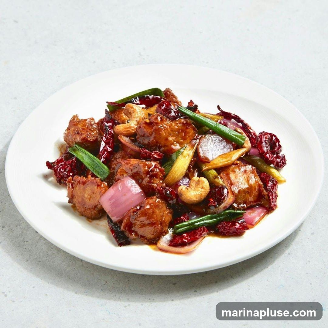 A white plate filled with a dish of spicy cashew chicken. The dish contains pieces of fried chicken coated in a dark, glossy sauce, mixed with cashews, red chili peppers, red onion chunks, and green onions. The plate is set on a light gray, textured surface.