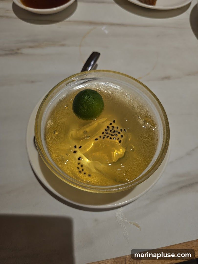 A close-up overhead view of a clear, golden dessert in a glass bowl. The dessert appears to be a jelly or panna cotta, with small black seeds suspended within it, resembling kiwi seeds. A whole, small green lime sits on top of the dessert. The bowl is placed on a white saucer, which rests on a marble-patterned tabletop. A spoon is visible behind the bowl. Other dishes are partially visible in the background.