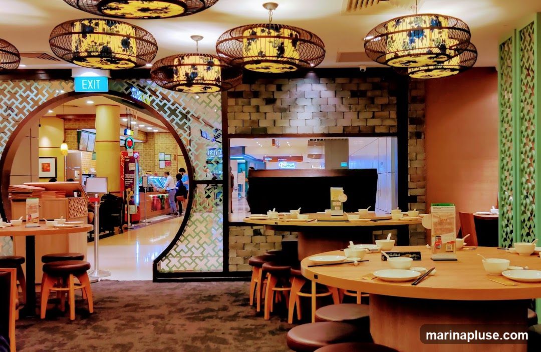 The image shows the interior of a restaurant with a distinctive Asian-inspired design. Several round tables are set with white bowls and plates, ready for diners. Overhead, large, decorative lamps with bamboo-like frames and floral patterns cast a warm glow. The walls feature a textured brick pattern and a decorative green screen. Through an arched entryway, a glimpse of another area is visible, including an "EXIT" sign and what appears to be a food counter with customers.