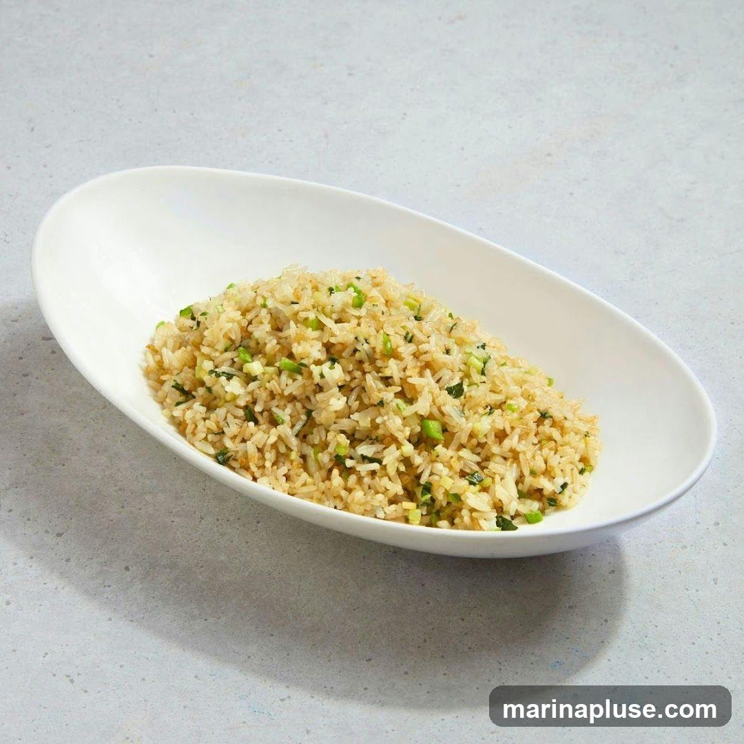A close-up shot of fried rice served in a white oval bowl. The rice is mixed with chopped green onions and other small green vegetables. The dish is presented on a light gray textured surface.