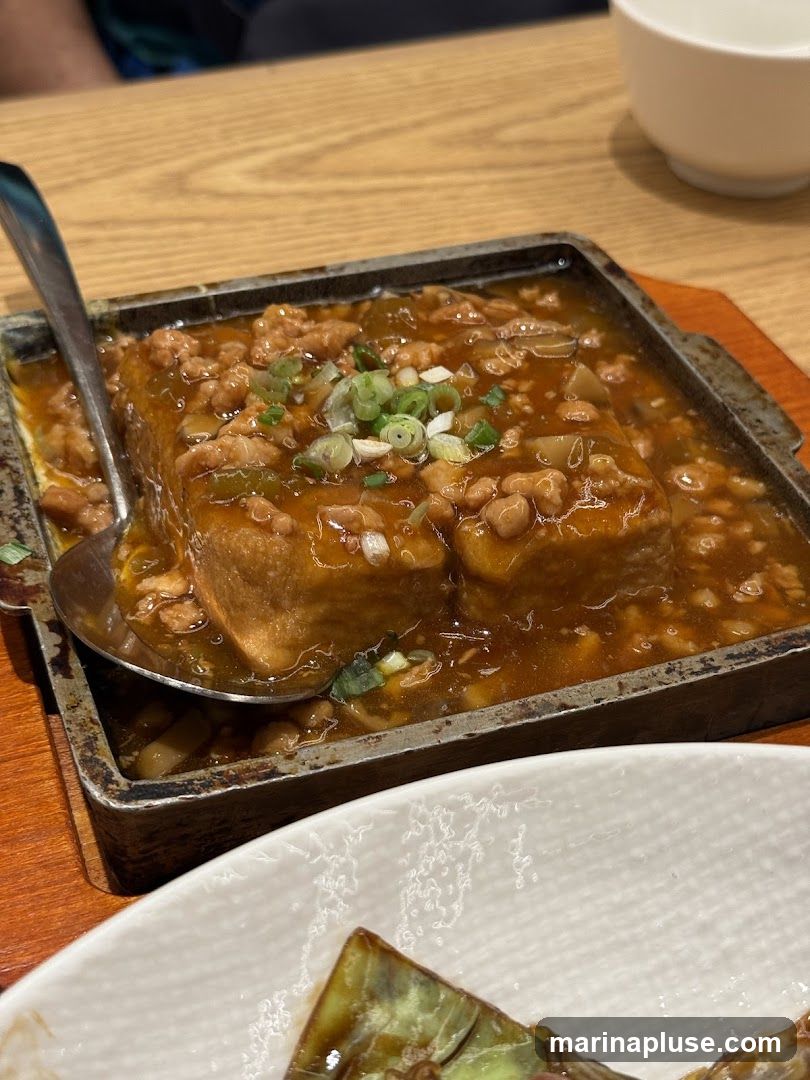 A close-up shot of a sizzling tofu dish served in a metal tray. The tofu is covered in a savory sauce with minced meat and garnished with chopped green onions. A spoon is placed in the tray, ready to serve. Another dish, possibly eggplant, is visible in the foreground. The meal is set on a wooden table, with a white bowl in the background.