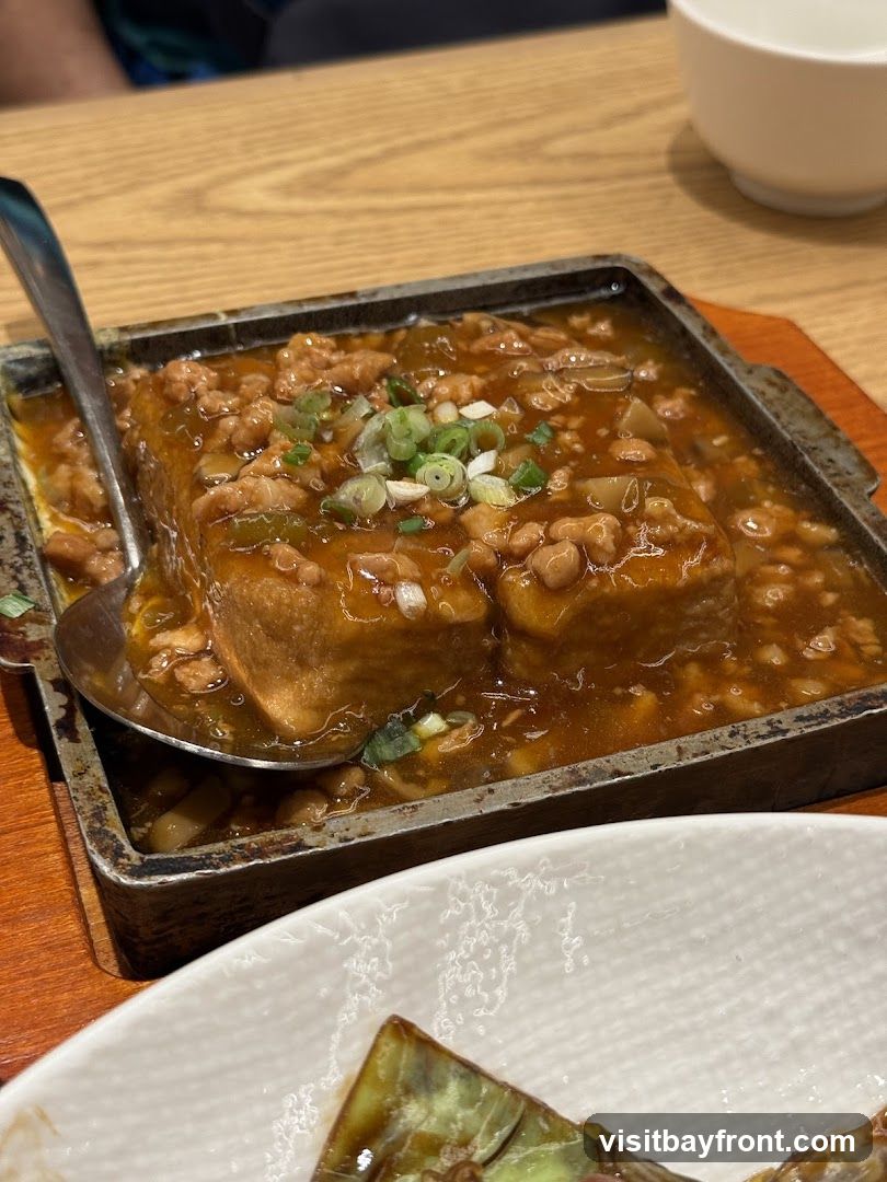 A close-up shot of a sizzling tofu dish served in a metal tray. The tofu is covered in a savory sauce with minced meat and garnished with chopped green onions. A spoon is placed in the tray, ready to serve. Another dish, possibly eggplant, is visible in the foreground. The meal is set on a wooden table, with a white bowl in the background.