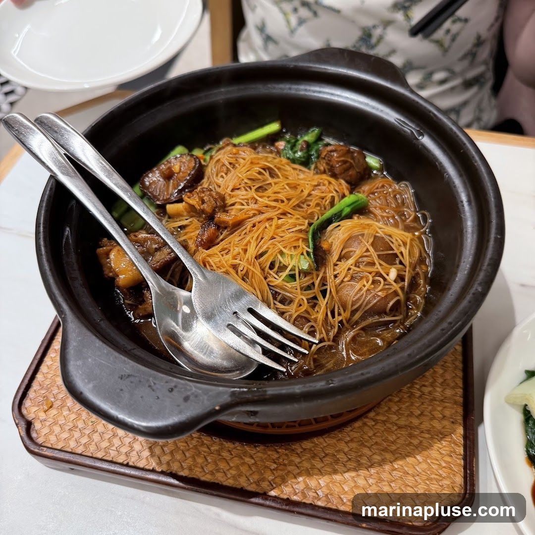 A close-up shot of a clay pot filled with braised noodles, chunks of meat, mushrooms, and green vegetables. A spoon and fork are resting on top of the noodles. The dish is served on a woven placemat.