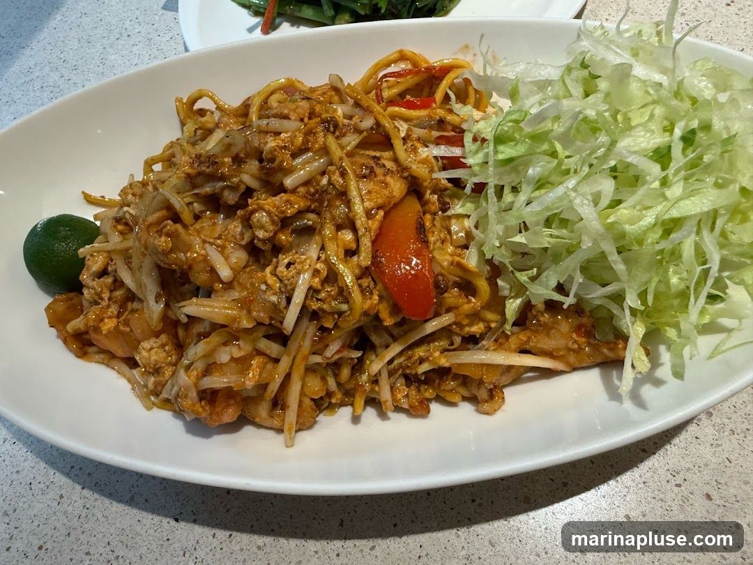 A close-up shot of a plate of noodles mixed with bean sprouts, scrambled eggs, and pieces of red bell pepper. A wedge of lime is on the left side of the plate, and a pile of shredded lettuce is on the right. Another plate with green vegetables is visible in the background.
