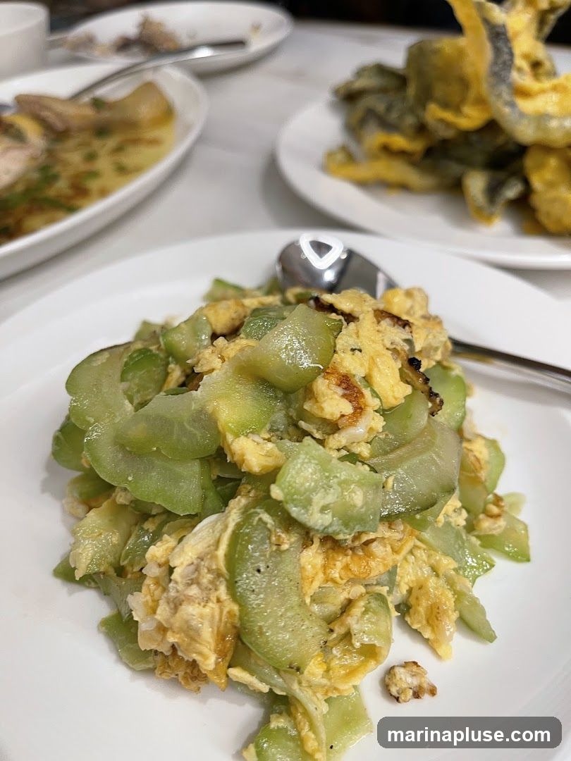 A close-up shot of a plate of stir-fried bitter melon with scrambled eggs. The dish is served on a white plate with a spoon. In the background, other dishes are visible on a table.